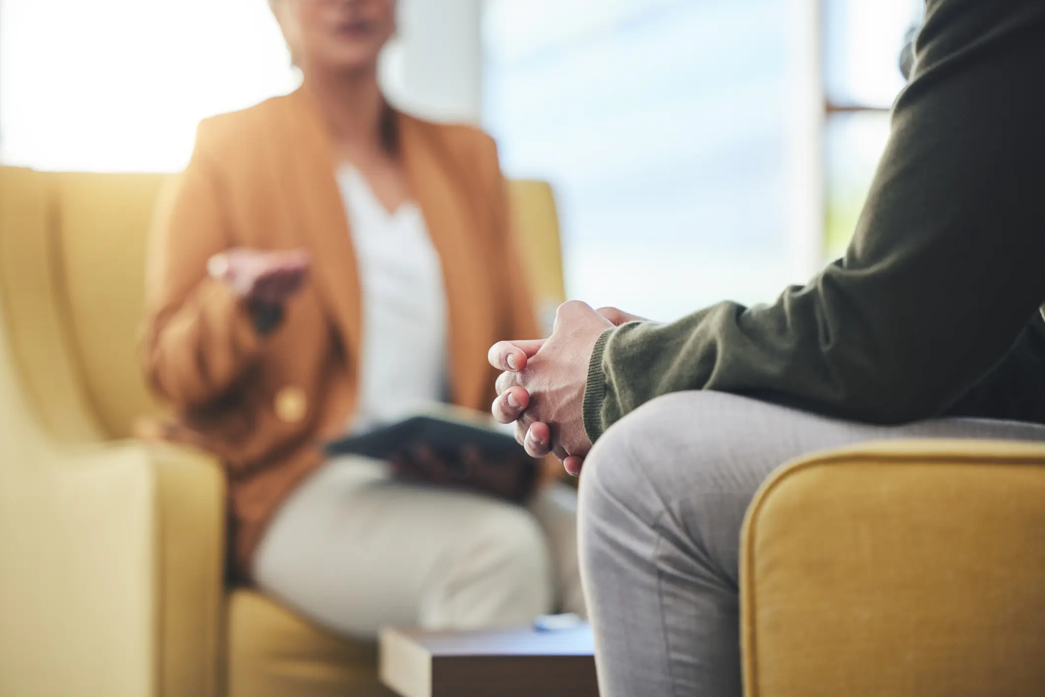 Man sitting with hands clasped on a chair across from a female counselor holding a tablet in a bright, calming therapy office—individual counseling session