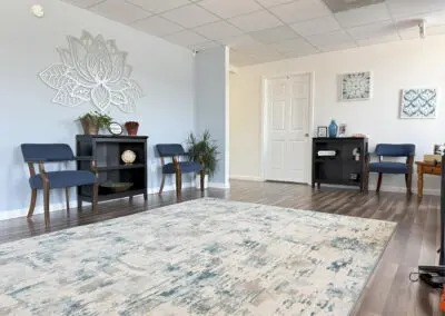 Waiting room with light blue walls, patterned area rug in foreground, four blue upholstered wooden chairs and two black shelving units holding plants, small decor and framed photos, lotus wall art above left shelf and framed artwork on right wall.