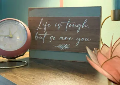 Wooden desktop sign reading "Life is tough, but so are you" sitting between a pink retro alarm clock on the left and a pink lotus candle holder on the right on a wood table surface