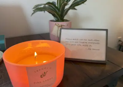 A lit pink "Pink Sands" soy wax candle on a wooden table beside a small potted plant and a framed Jim Henson quote about watching out for each other and enjoying life