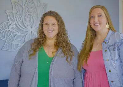Two women standing side-by-side in a counseling office; one wears a green top and gray cardigan with curly hair, the other wears a pink dress and denim jacket with long straight hair, a large decorative lotus wall art behind them.