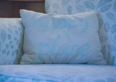 Decorative rectangular throw pillow with pale blue embroidered script reading "happy place" resting on a light-patterned armchair upholstery