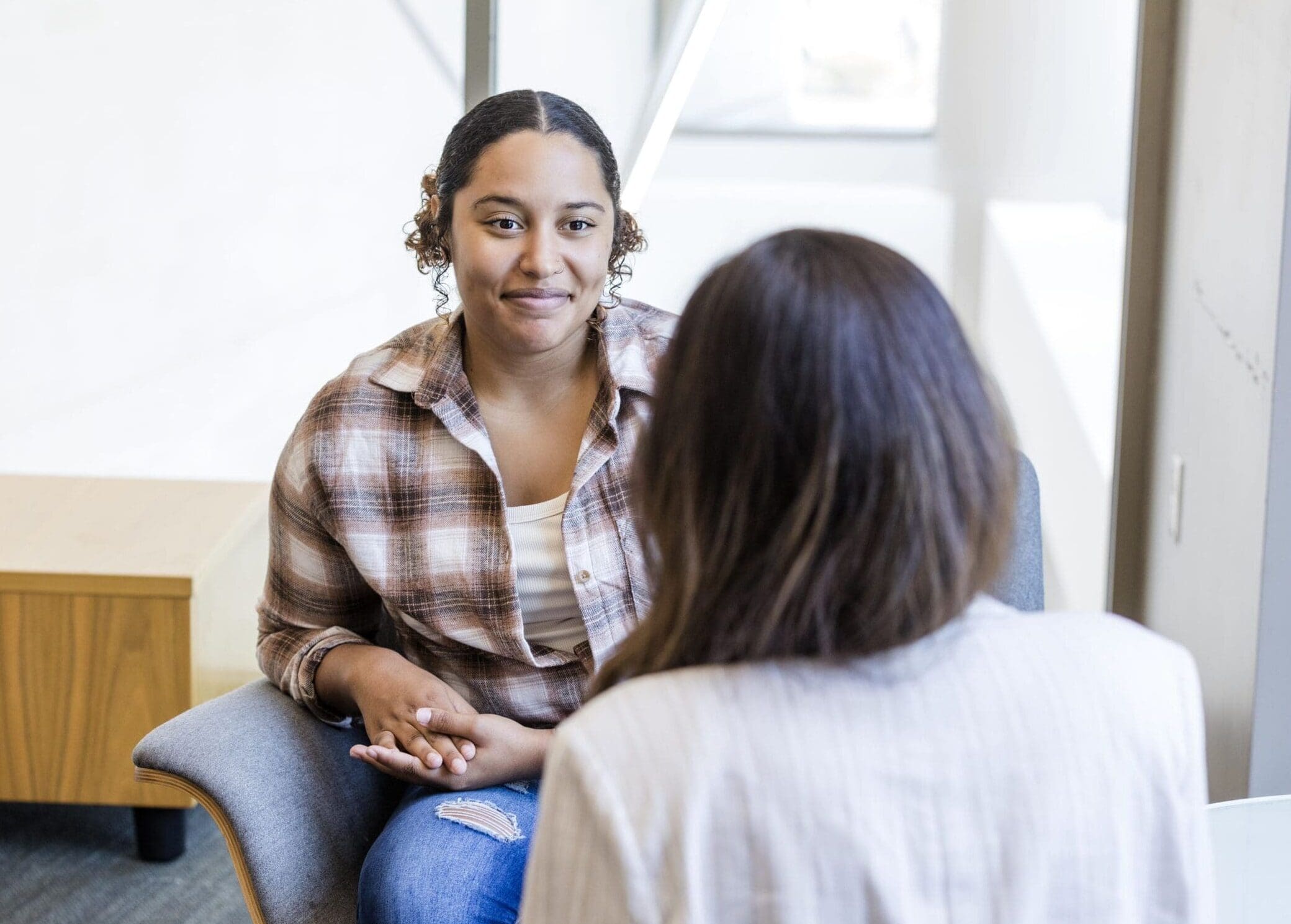 Two women sitting in a bright counseling office during a therapy session, one wearing a plaid shirt and jeans with hands clasped, the other facing her from a chair, conveying a calm, supportive counseling setting for mental health and patient resources